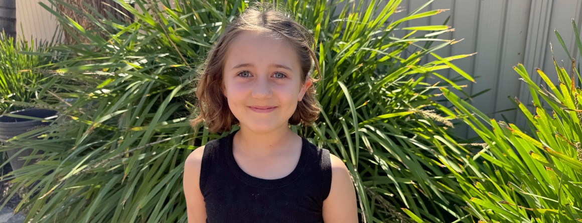 A primary-school aged girl smiles to the camera. She has short, light brown hair and stands in front of a bright green plant.